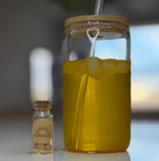 Glass jar with yellow liquid and ice cubes, next to a small bottle on a reflective surface.