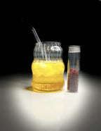Clear container with yellow liquid and black straw next to a small container with red and white objects on a dark background