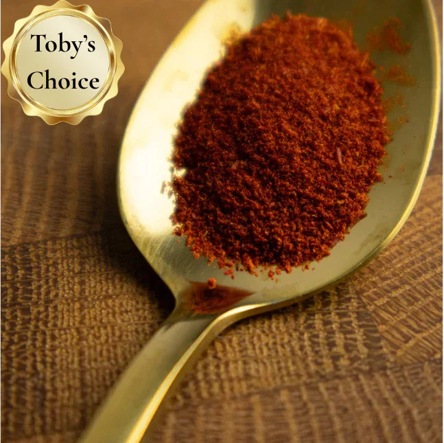 Gold spoon filled with red spice powder on a textured brown background, with 'Toby's Choice' logo